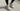 Woman wearing HOKA Clifton 10 Putty Grout running shoes with black leggings while standing on a concrete sidewalk, showcasing the neutral beige colorway and cushioned performance design.