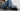 Woman wearing black UGG Tasman II slippers with relaxed blue jeans and gray socks, featuring signature embroidered trim and suede uppers.